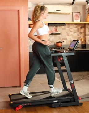 Uma mulher em traje esportivo caminha em uma esteira na cozinha, segurando um celular. A esteira tem uma tela, e a cozinha é moderna e organizada.