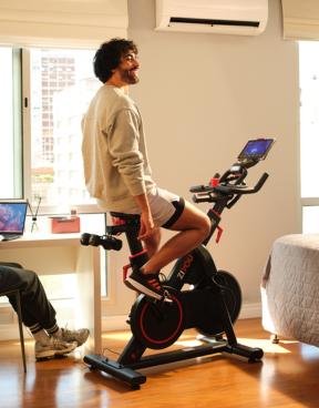 Uma pessoa se exercita em uma bicicleta ergométrica em uma sala iluminada, com um laptop em uma mesa próxima e uma cama ao fundo.